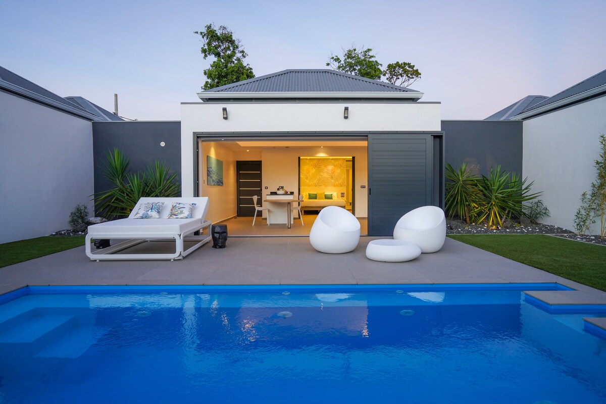 A modern outdoor space is captured, featuring a clear blue swimming pool reflecting the sky. Comfortable white lounge chairs are positioned beside the pool, offering a relaxing spot. The interior space, visible through large sliding doors, is well lit and decorated with contemporary artwork.