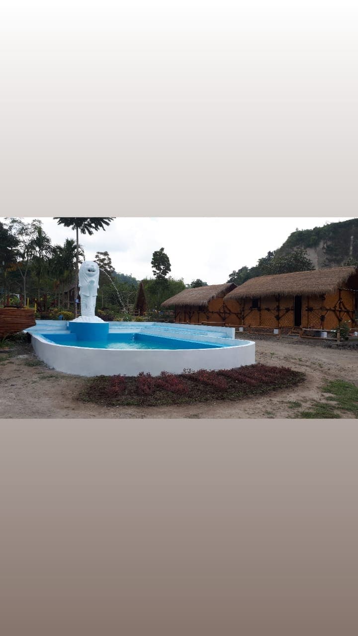 An outdoor swimming pool features a blue surface with a fountain at its center. Surrounding the pool, rustic wooden structures are visible, blending with the natural environment. Lush greenery and sandy areas are present, contributing to the tranquil setting.