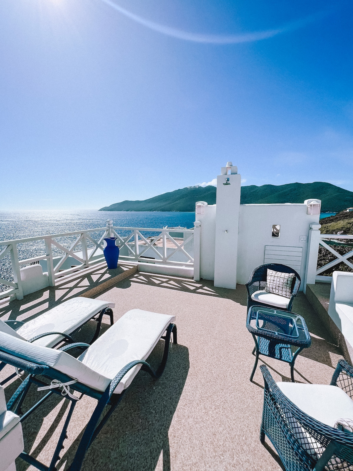 An outdoor terrace features a stunning ocean view, with two lounge chairs and a small table arranged for relaxation. The white railing contrasts with the blue sea and mountains in the background, enhancing the serene atmosphere of the space.