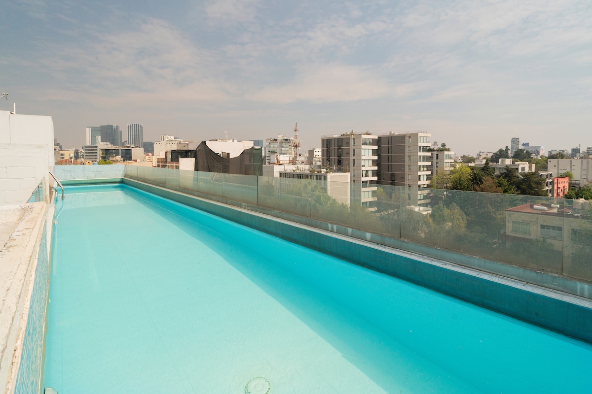An expansive rooftop pool is surrounded by clear glass railings, offering unobstructed views of the city skyline. The bright blue water contrasts with the surrounding modern buildings and distant landmarks under a clear sky.