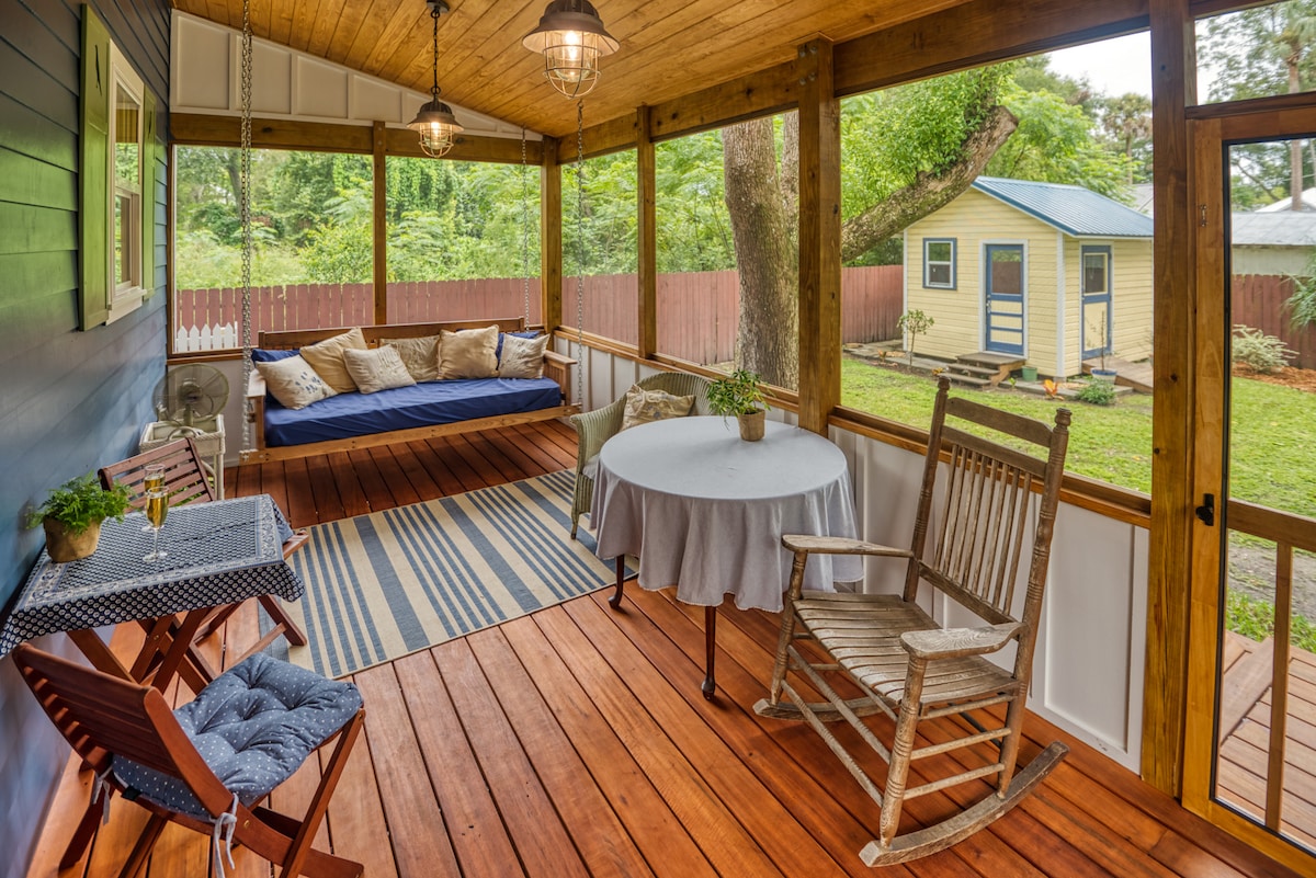 A spacious screened-in porch features a hanging daybed, rustic table, and two chairs. The wooden floor is complemented by a striped rug. Greenery is visible through the screens, with a distant view of a small shed and a fenced yard beyond.