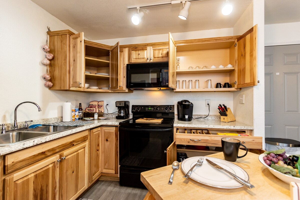 A well-equipped kitchen features wooden cabinetry, a black stove, and a microwave. Granite countertops provide ample space for meal preparation. Dining essentials, including plates and cutlery, are neatly arranged on a wooden table alongside a bowl of fresh fruit.