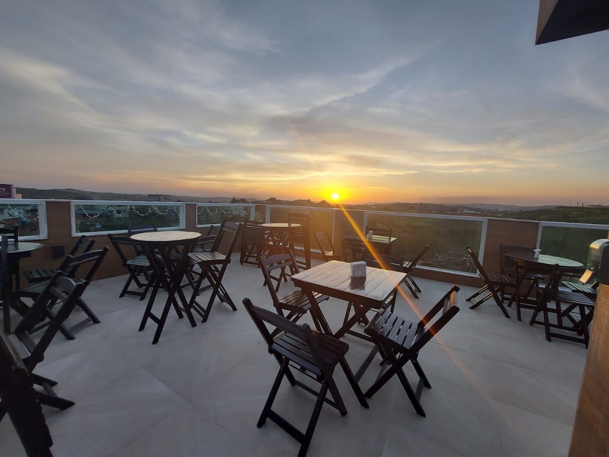 A rooftop terrace is equipped with several wooden tables and chairs, arranged to provide views of the sunset. Soft light illuminates the space, highlighting the clean lines of the furniture against the expansive sky.
