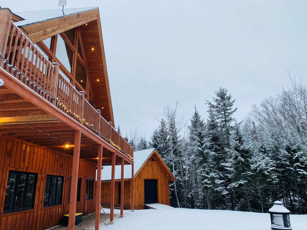 The exterior of a Scandinavian-style wooden chalet is captured amidst a snowy landscape. A spacious deck wraps around the upper floor, with large windows reflecting the serene winter scenery. Surrounding evergreen trees are dusted with snow, adding to the tranquil atmosphere.