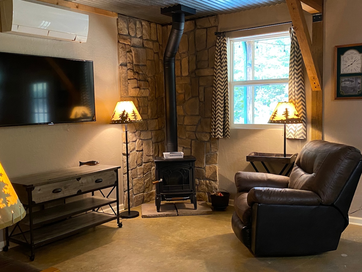 A cozy living area features a stone accent wall with a modern TV mounted above a wooden media console. Two standing lamps provide soft lighting beside a plush leather recliner. A compact wood stove adds warmth, while a window allows natural light to fill the space.