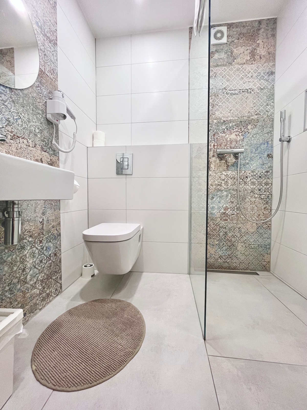 A modern bathroom is showcased, featuring a walk-in shower with a glass partition and a rain showerhead. A wall-mounted mirror is above a compact sink, while a toilet is positioned beside it. Textured wall tiles add visual interest to the space.