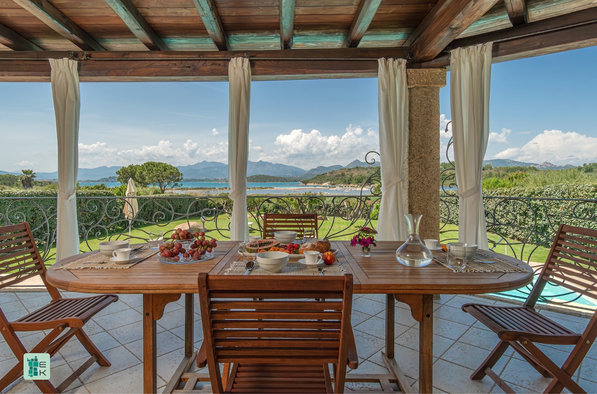 An outdoor dining area is framed by lush greenery and scenic views of the landscape. A wooden table is set with plates of fresh fruits and pastries. Light, flowing curtains provide a gentle privacy while allowing natural light to fill the space.