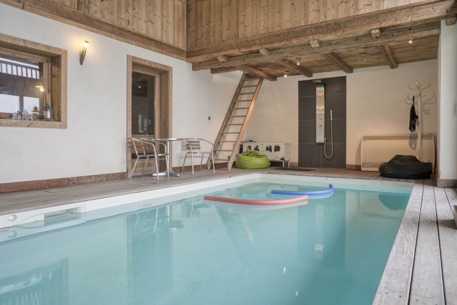 An indoor heated pool is visible, surrounded by wooden walls and beams. Two colorful floats are placed on the water's surface. A staircase and a shower area are present in the background. Nearby, small tables and chairs are arranged for relaxation.