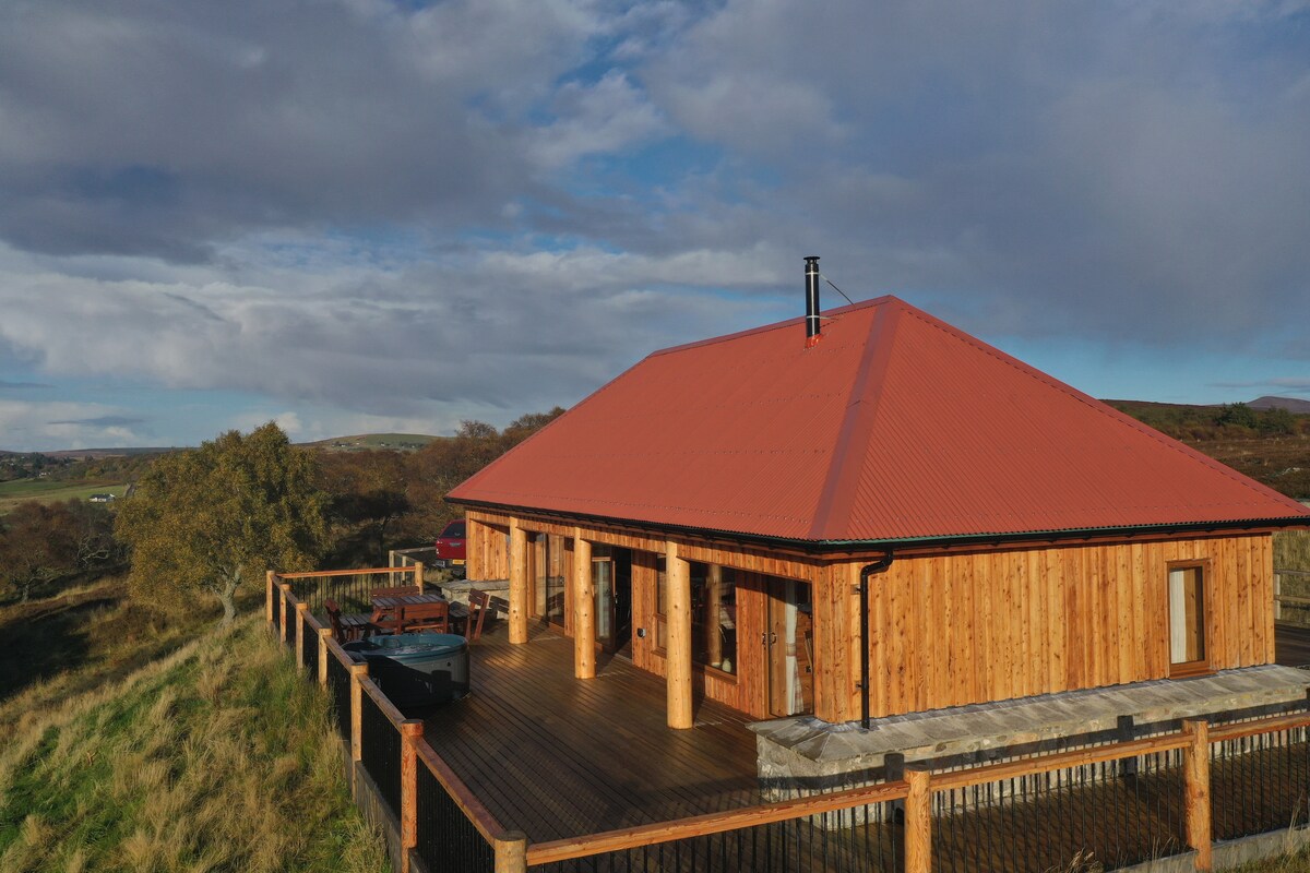 A spacious wooden lodge with a crimson roof is featured, situated on elevated ground. Surrounding the lodge is a wooden deck, equipped with seating and a hot tub, while sweeping views of the expansive countryside and hills are visible in the background.
