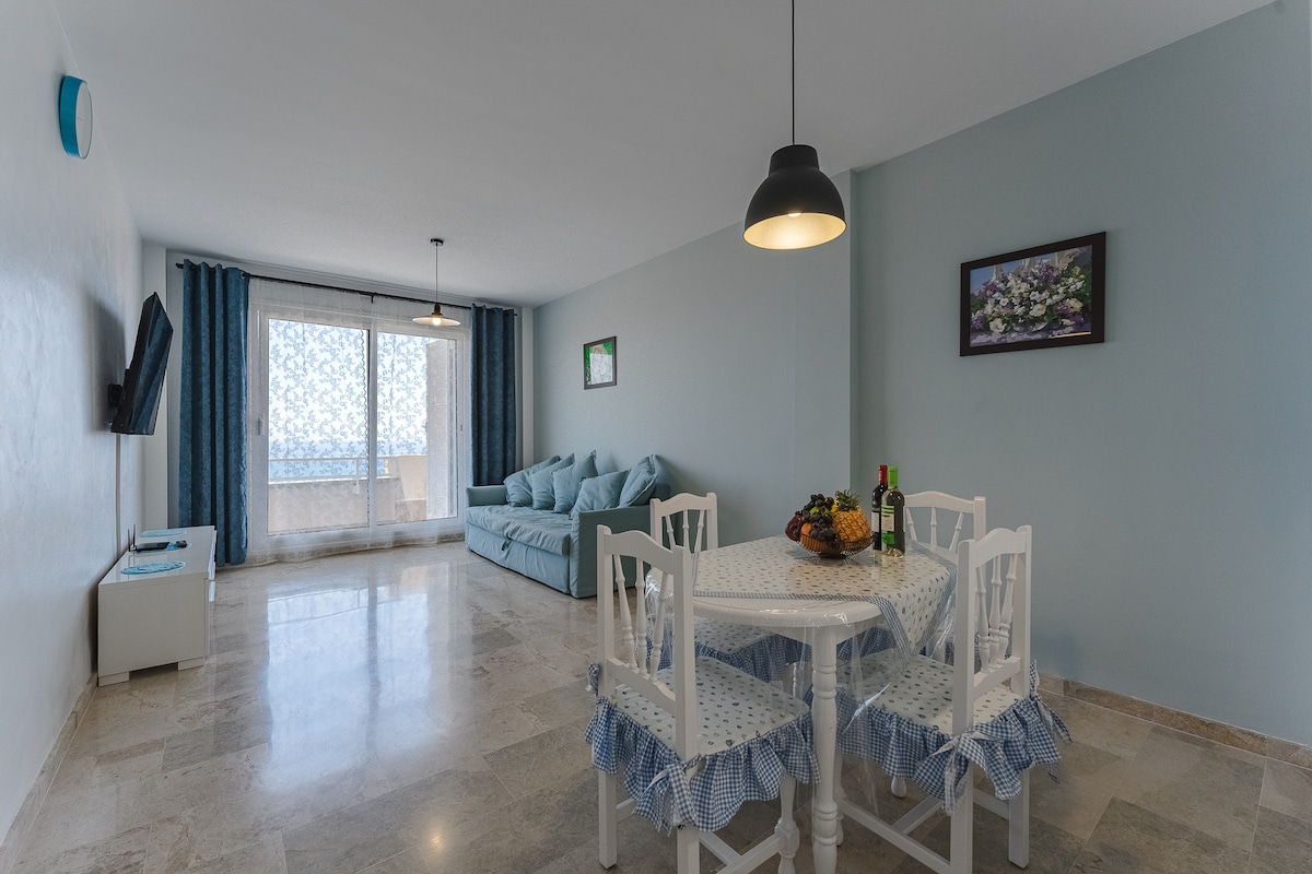 A well-lit living area features a light blue seating arrangement and a white round dining table with blue and white patterned chair covers. Large glass doors provide access to a balcony, while marble flooring adds an elegant touch. A framed floral print decorates the wall.