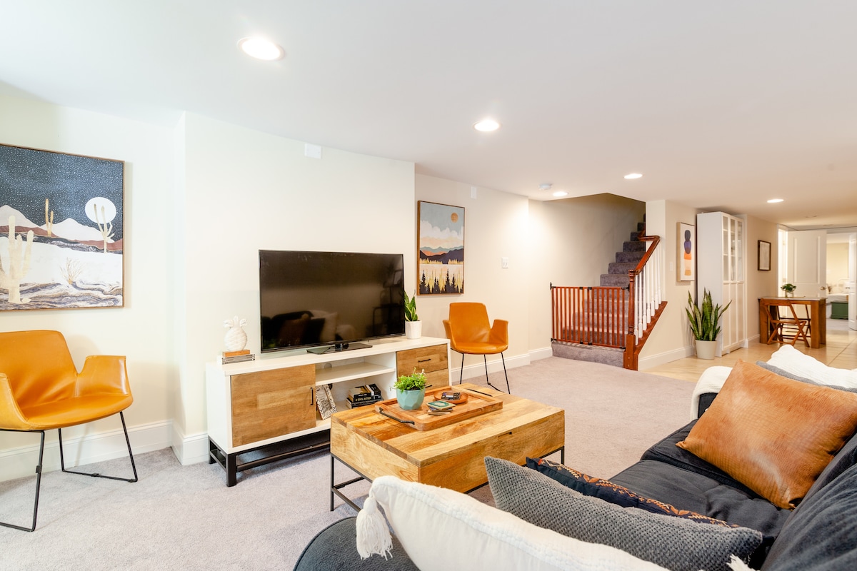 A spacious living area is furnished with a large sectional couch and a rustic wooden coffee table. Two modern orange chairs are positioned beside a television on a media console. Light-colored walls and soft carpet create a welcoming environment, complemented by decorative artworks and plants.