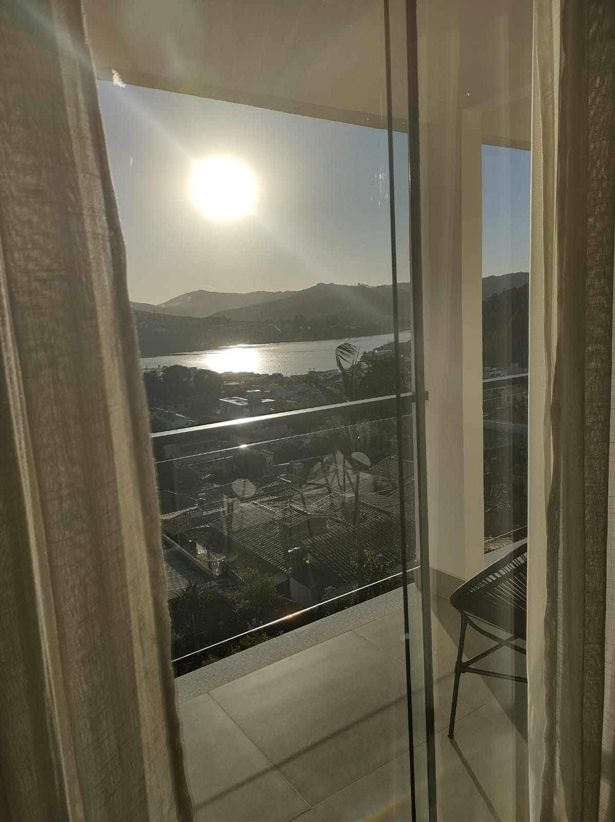 Soft sunlight filters through sheer curtains, illuminating a modern balcony. A simple chair is positioned on the balcony, offering a direct view of the lake and surrounding hills in the distance, enhancing the serene setting.