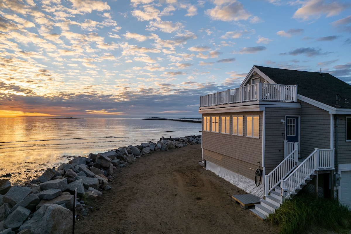 The oceanfront house is framed by a sandy beach and rocky shoreline. Soft hues of sunset illuminate the sky, reflecting on the calm water. The exterior features a light gray facade, with a staircase leading to a spacious deck overlooking the scenic view.