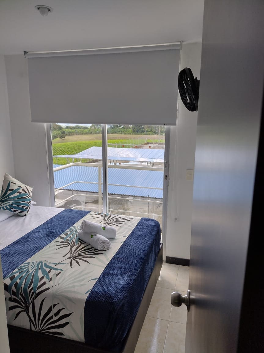 A bright bedroom is featured, with a spacious bed dressed in a patterned coverlet. Natural light enters through a large window that offers views of lush greenery. A fan is positioned nearby for comfort, and a soft towel is neatly arranged on the bed.