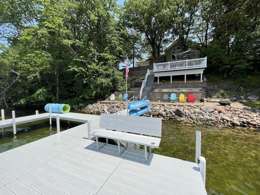 A boat dock extends into the clear water, featuring a white bench and colorful chairs positioned along the shoreline. Lush greenery surrounds the area, while a two-level deck with additional seating is visible in the background, enhancing the outdoor leisure space.