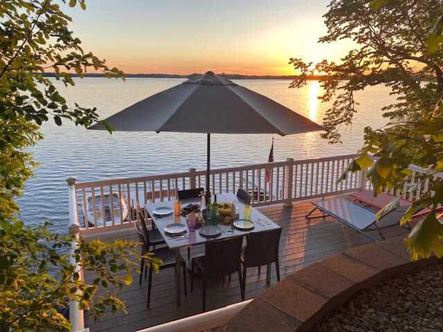 A dining area is presented on a deck overlooking the lake, featuring a table set for meals under a large umbrella. The serene scene is enhanced by a setting sun casting warm light over the water, framed by leafy trees.