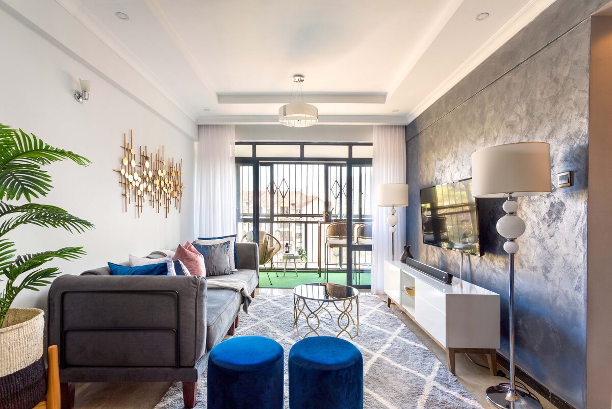 A modern living area is showcased with a plush sofa accompanied by pastel cushions. A glass coffee table sits on a textured area rug, and a wall-mounted television is present. Large sliding doors lead to a balcony, while floor lamps and decorative wall art enhance the space.