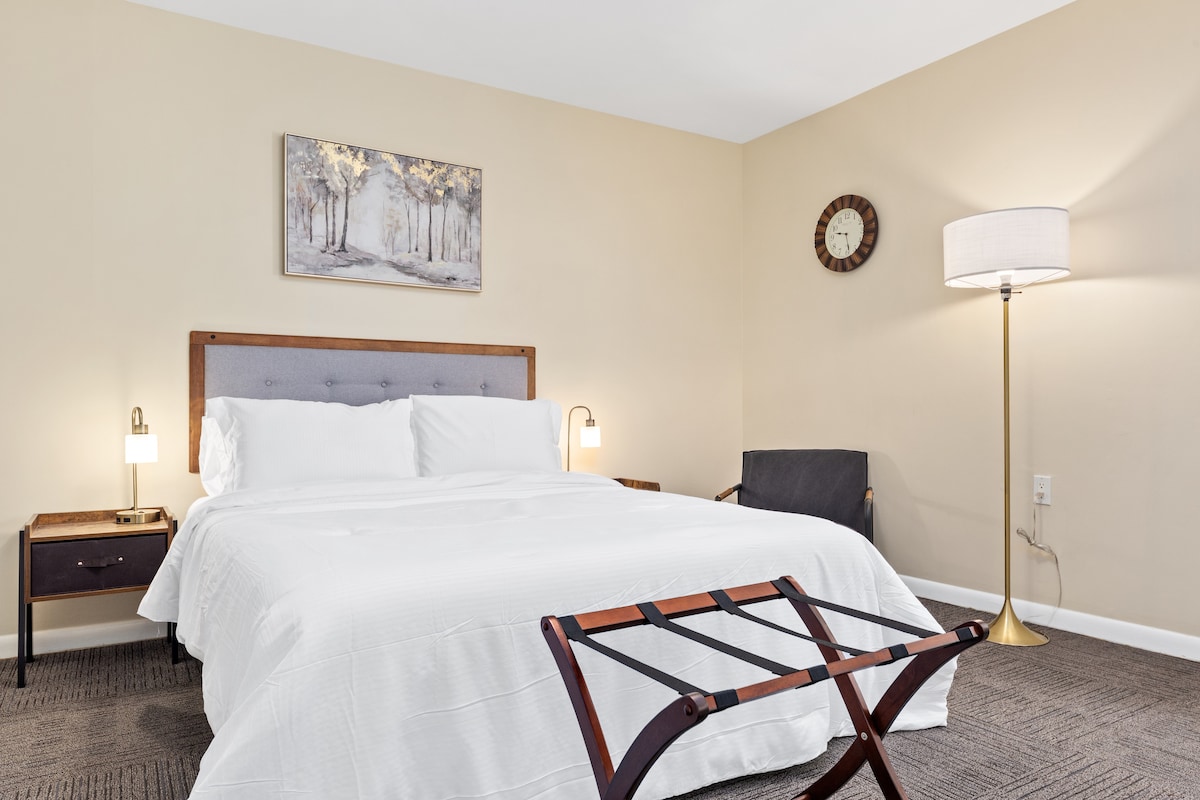 A well-appointed sleeping area showcases a spacious bed dressed in crisp white linens. Soft lighting emanates from bedside lamps, illuminating the neutral-colored walls. A decorative piece of art hangs above the bed, and a modern chair and lamp are positioned in the corner.