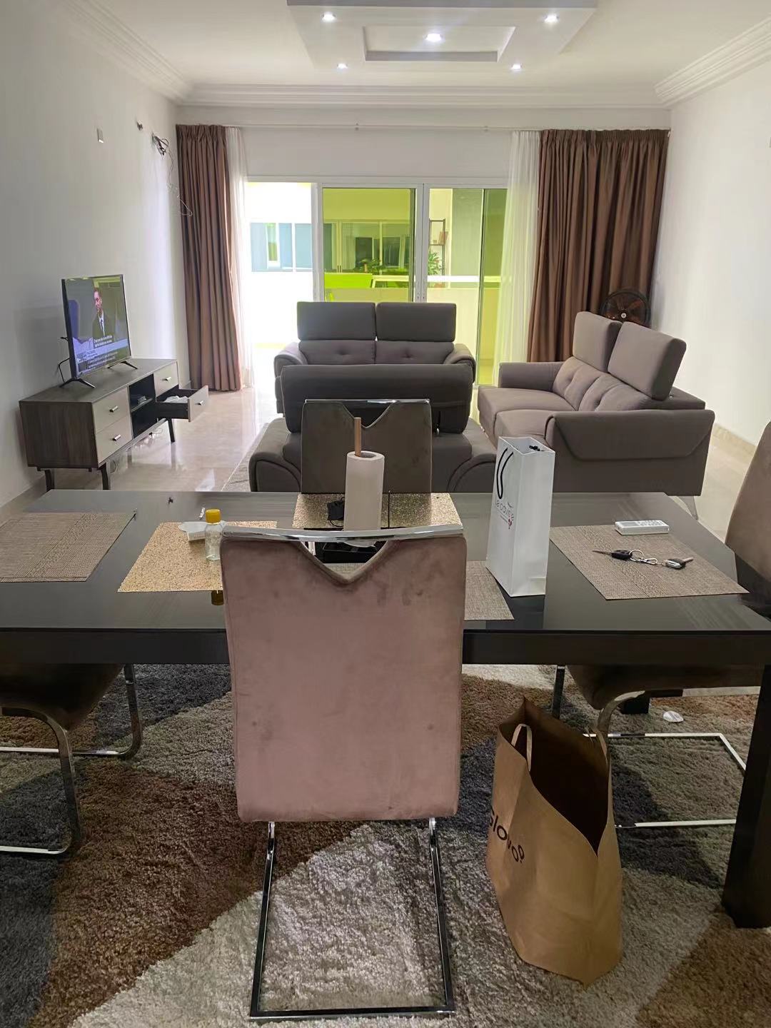 A spacious living area features a comfortable sofa set facing a television. A dining table is set for meals, accompanied by elegant chairs. Large windows allow natural light to fill the room, while light curtains frame the view of the outside.