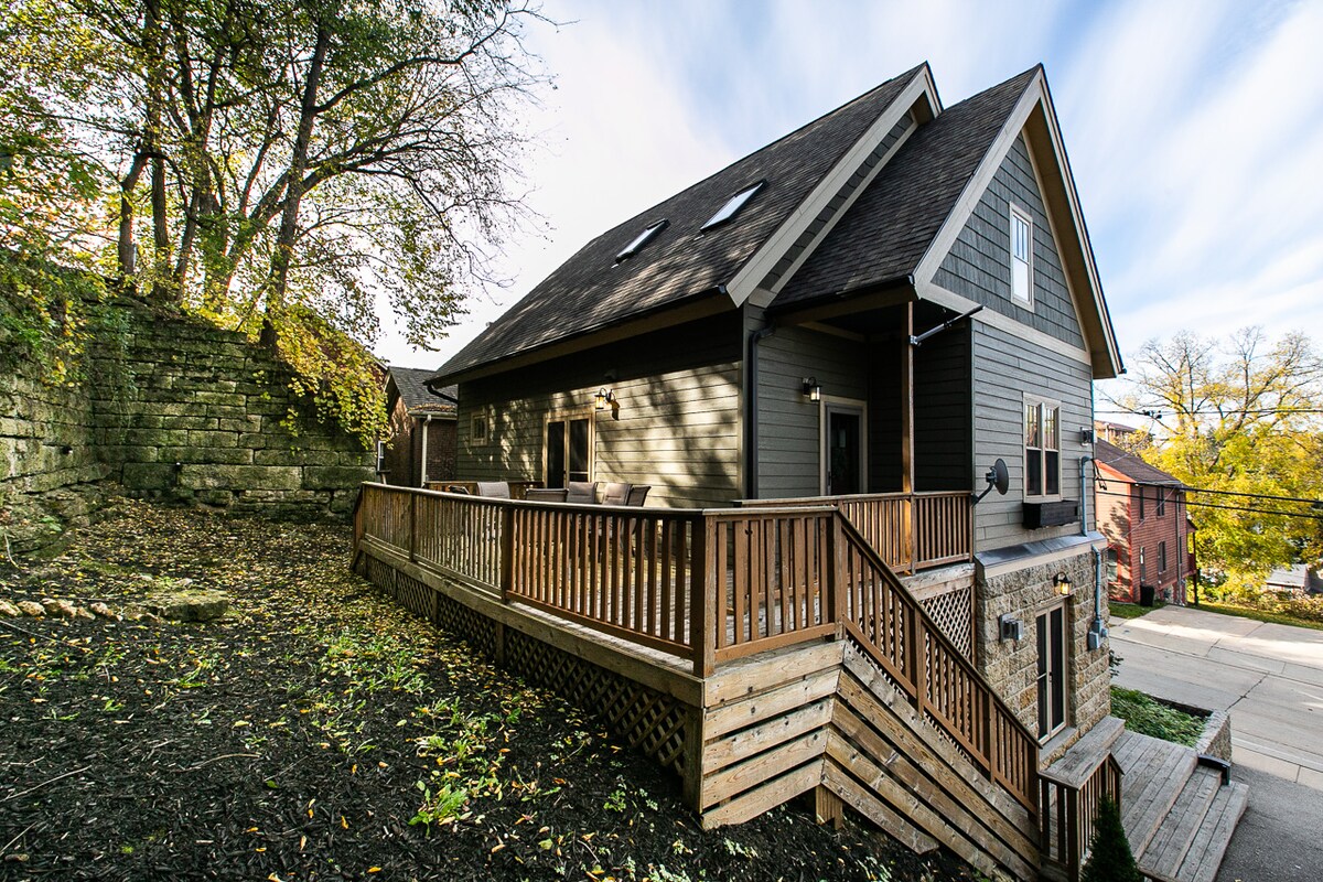 The exterior of The Arlington Cottage is shown, featuring a wooden deck with a railing that extends along the front. The structure, with its gray siding and sloping roof, is surrounded by greenery and stone landscaping. A pathway leads from the deck to the surrounding area.