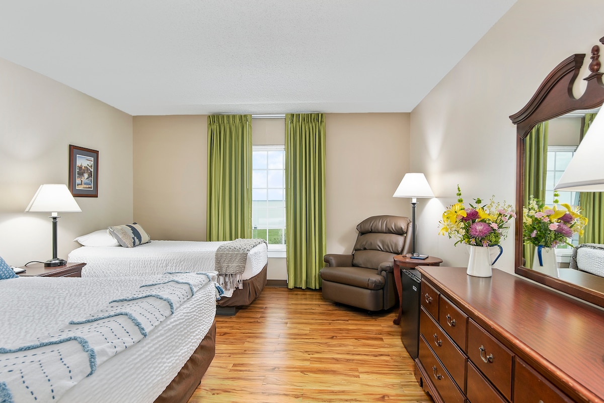 A room featuring two beds, one queen and one extra-long twin, positioned near a window with green curtains. A lounge chair recliner is placed in the corner, complemented by a dresser and mirror. Natural light enhances the wood-like flooring throughout the space.