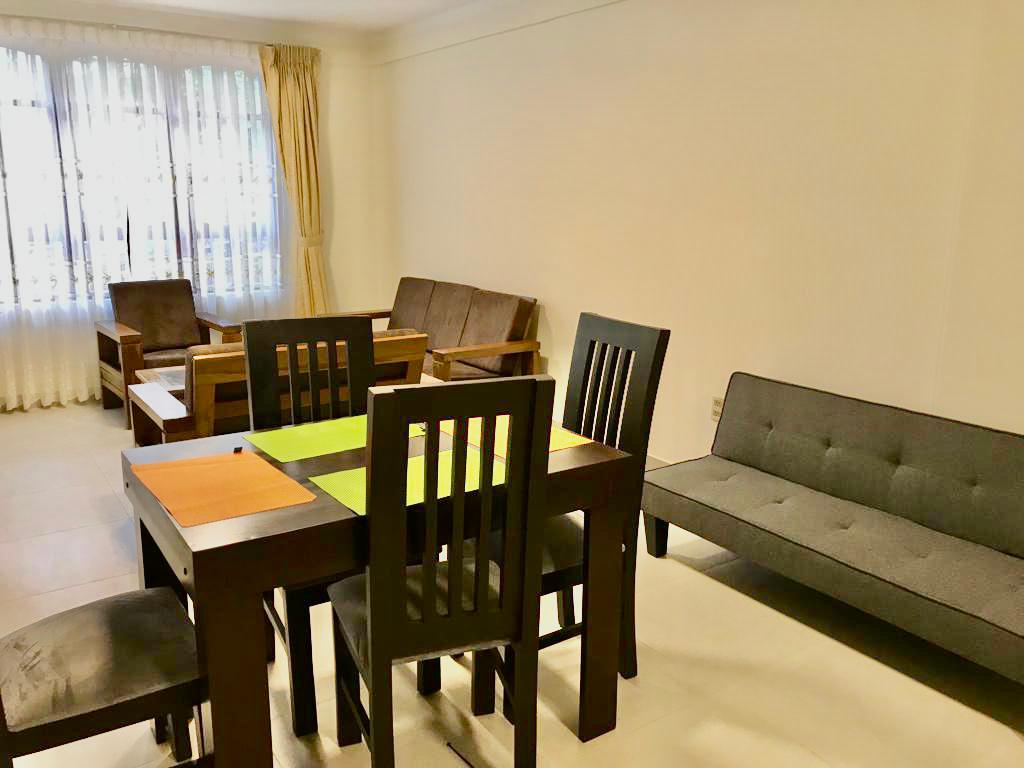 A spacious living area is displayed, featuring a dark dining table surrounded by five matching chairs. An additional seating area includes two upholstered couches and a wooden chair. Natural light filters through large windows with sheer curtains, enhancing the room's bright and airy feel.