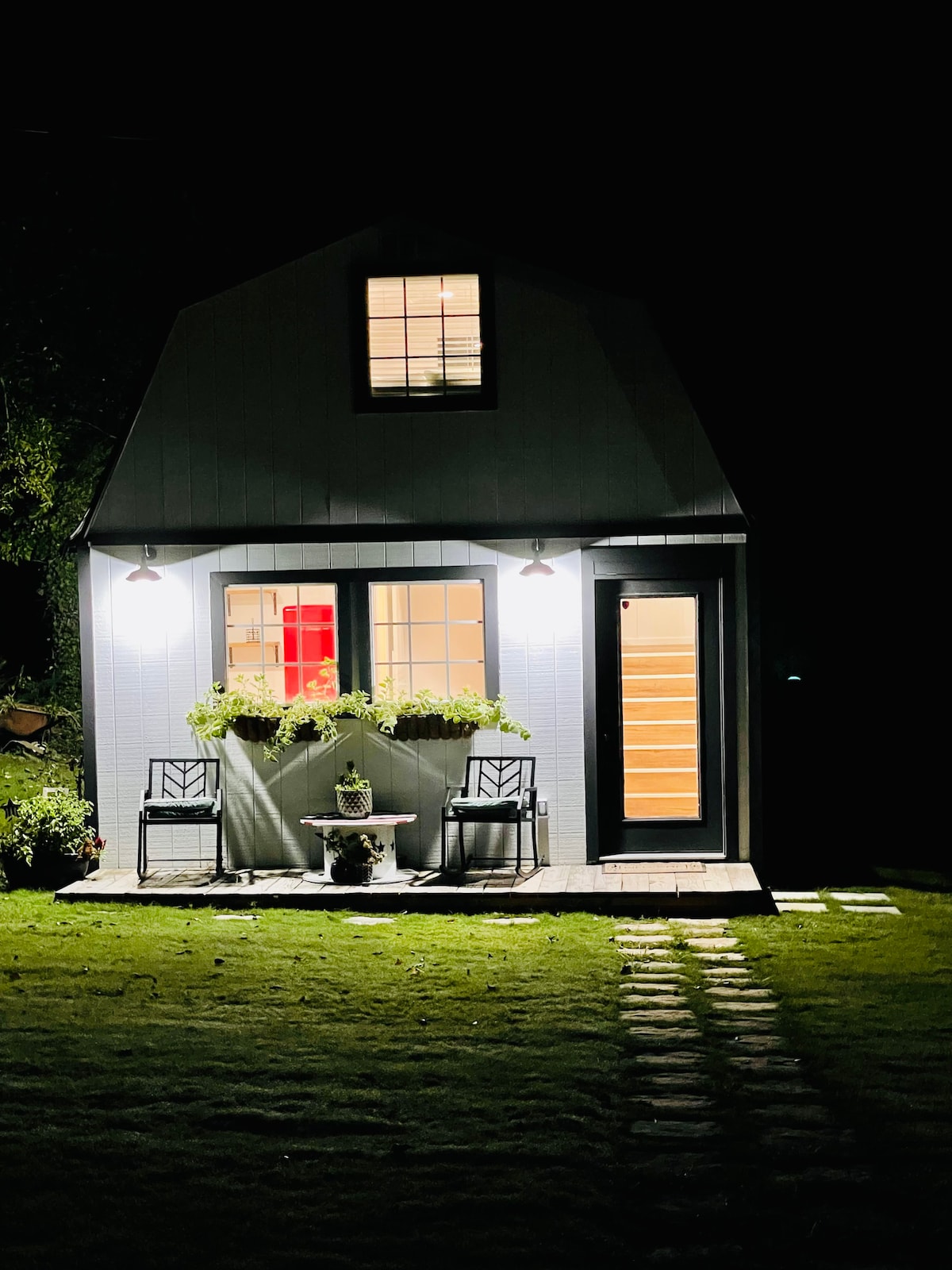 The cottage exterior is illuminated at night, featuring two chairs on either side of a small table. A flower box with greenery enhances the front windows, while a pathway composed of stone slabs leads toward the entrance. Soft lighting highlights the architectural details.