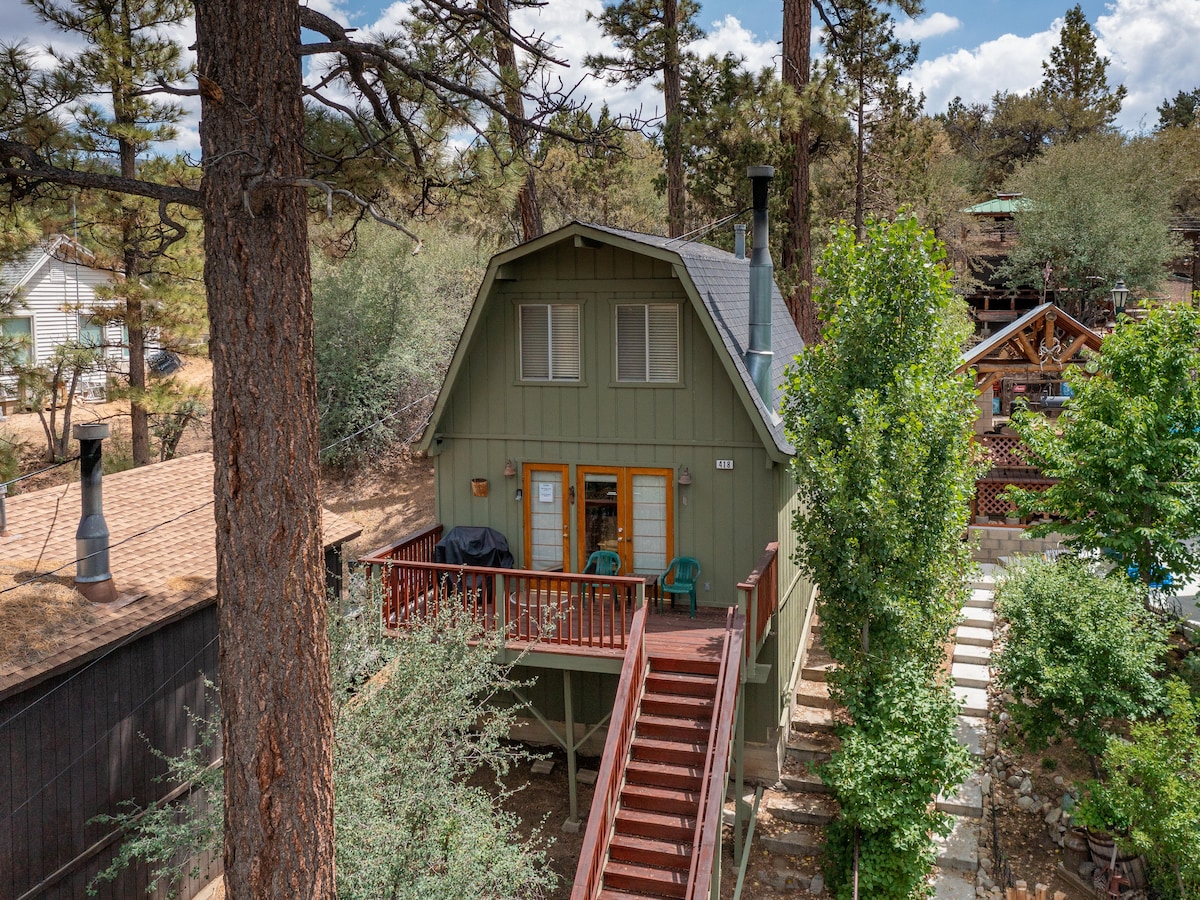 The exterior of the cabin features a green facade with a pitched roof. A spacious front deck is adorned with two chairs, and steps lead down from the deck. Surrounding foliage includes tall trees, enhancing the serene mountain setting.