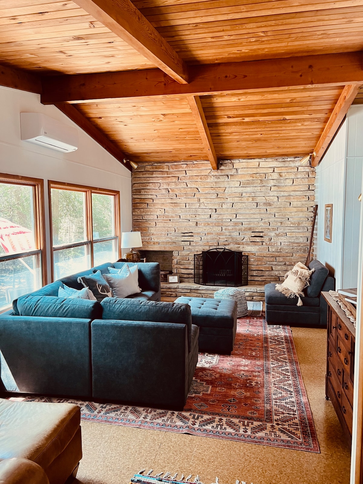 A cozy living area features a stone fireplace, surrounded by large windows that invite natural light. Comfortable seating arrangements include a dark sectional sofa with accent cushions, complemented by a patterned area rug. A wooden cabinet adds a warm touch to the space.