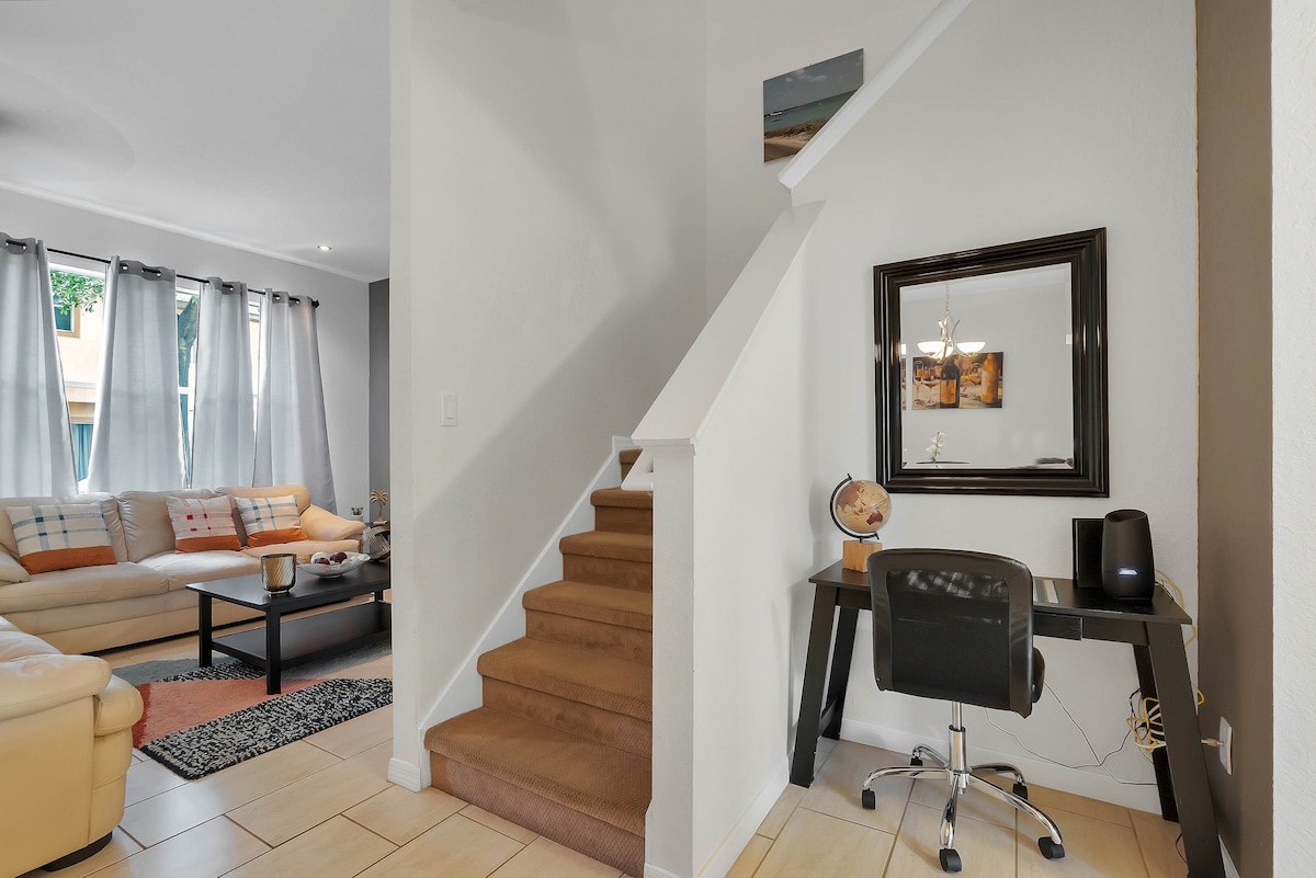The living area features a light-filled space with large windows adorned by grey curtains. A comfortable sectional sofa and an area rug are visible. A dedicated workspace is set up near the staircase, equipped with a desk and a chair, creating a functional corner.