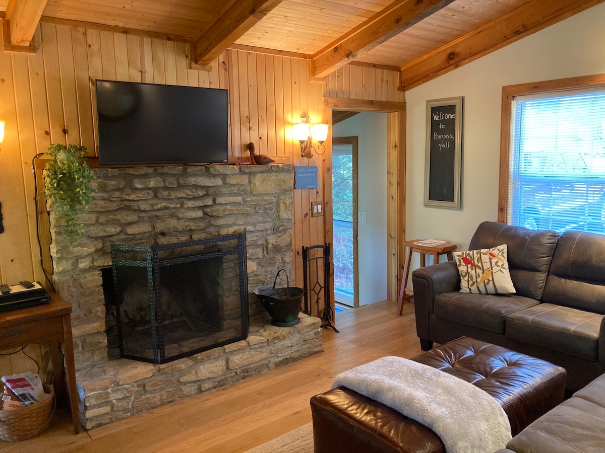 The living room features a stone fireplace with an iron grate and a mounted flat-screen TV above it. Warm wooden walls complement a leather sofa and an ottoman, with natural light filtering through a nearby window.