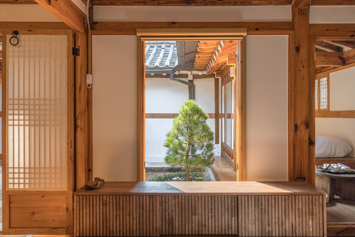 A wooden bench runs along a spacious room, providing a clear view of a small pine tree in the courtyard. Light enters through an open door, highlighting the traditional wooden beams and minimalist design elements of the interior space.
