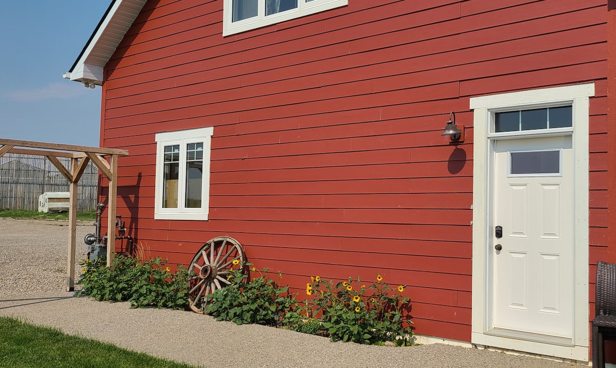 A charming red exterior of the guest suite is complemented by a landscaped pathway. Bright sunflowers and a rustic wagon wheel add a touch of character. The entrance is marked by a white door and windows, providing natural light and an inviting entry.
