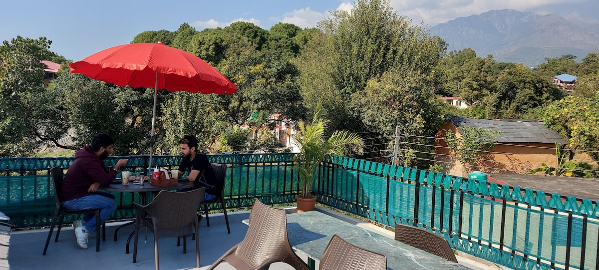 A rooftop terrace is equipped with a large red umbrella, offering shade over a table where two guests are seated. Surrounding greenery and distant mountains create a peaceful backdrop, while the area is furnished with several brown chairs and a clear blue sky overhead.