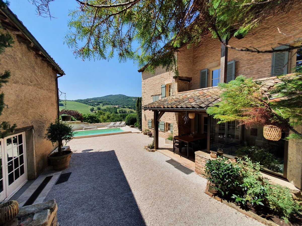 A sunlit courtyard is framed by the charming architecture of La Maison des Iris. A private pool is visible in the background, surrounded by lush greenery. The area is enhanced by a covered dining space, providing an inviting spot for outdoor meals and relaxation.