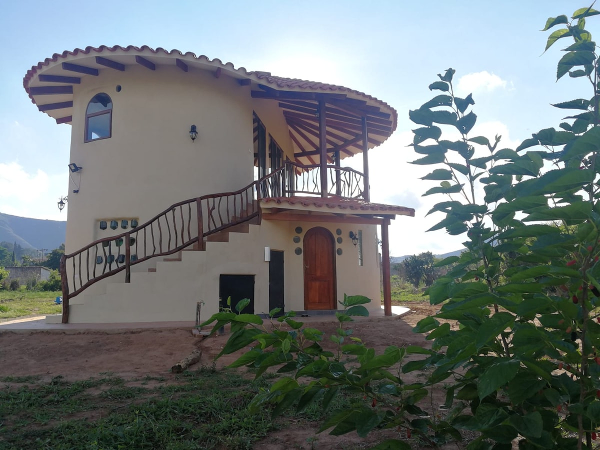 A circular rustic cabin is showcased, featuring a curved staircase leading to the entrance. Large windows create an open ambience, allowing views of the surrounding landscape. A natural setting complements the structure, with greenery and open space visible in the foreground.
