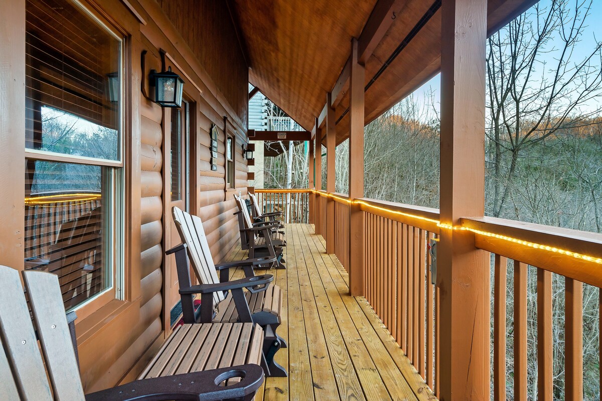 A spacious wrap-around deck features wooden gliders and a sunlit pathway. The log cabin structure is complemented by wooden railings, with trees and natural scenery visible beyond. The peaceful setting invites relaxation in the fresh air.