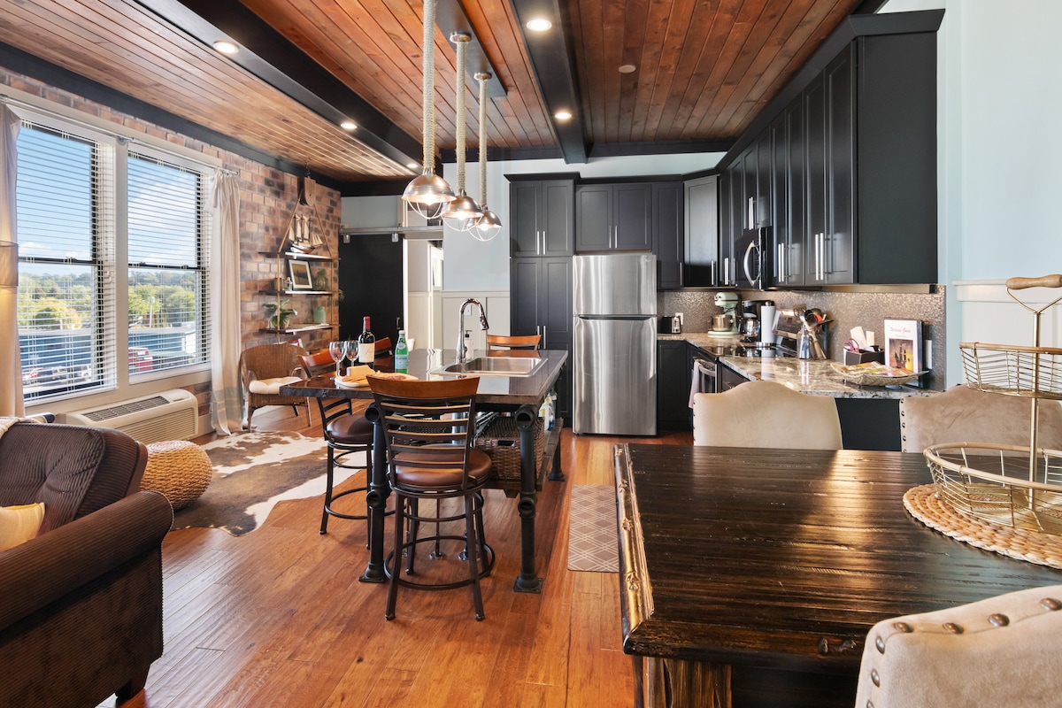 An open-concept area is highlighted by a mix of wooden beams and sleek cabinetry. A spacious kitchen with a large island is visible, along with a dining table and cozy seating arrangements. Natural light fills the room through the large windows, enhancing the inviting atmosphere.