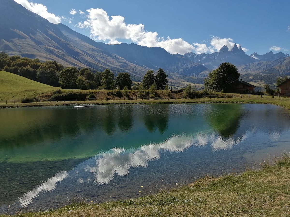 A serene pond reflects the blue sky and surrounding mountains, creating a harmonious connection with nature. Lush green fields frame the water's edge, while trees provide a backdrop of natural beauty. The distant peaks enhance the tranquil atmosphere of the landscape.