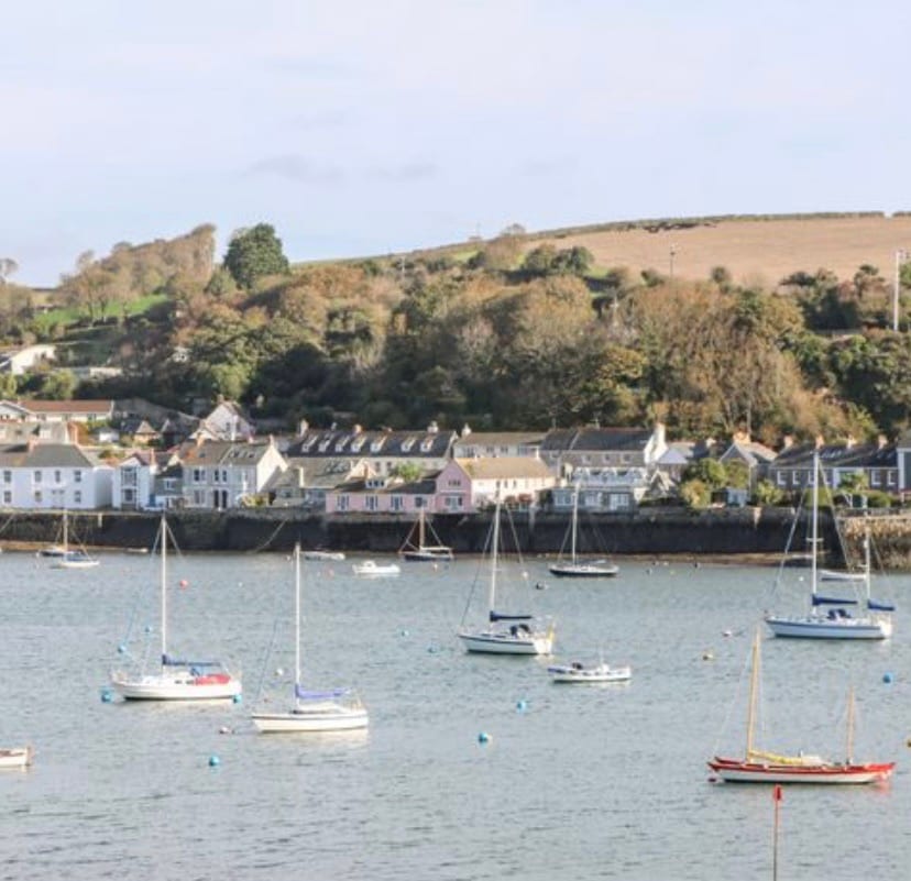 A view of a calm harbor filled with several sailboats anchored in the water. The shoreline features a collection of charming buildings, including a mix of colorful facades. Lush green hills are visible in the background, adding depth to the scenery.