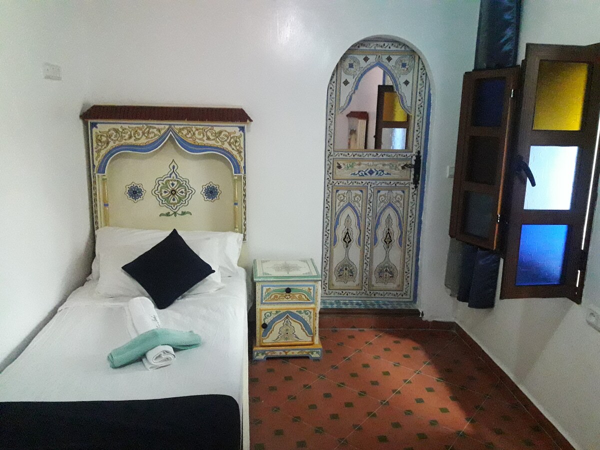 A single bed with neutral bedding is positioned against a decorative wall. An intricately designed door leads to the exterior, while a colorful bedside table adds charm. Natural light filters through the partially open windows, illuminating the terracotta floor tiles.