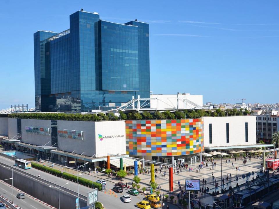 A large shopping center is showcased with a modern design. The exterior features a colorful, mosaic-like facade and expansive glass windows. Surrounding green landscape and pathways lead to the entrance, where pedestrians are visible. Towering above is a high-rise building with blue glass reflecting the sky.