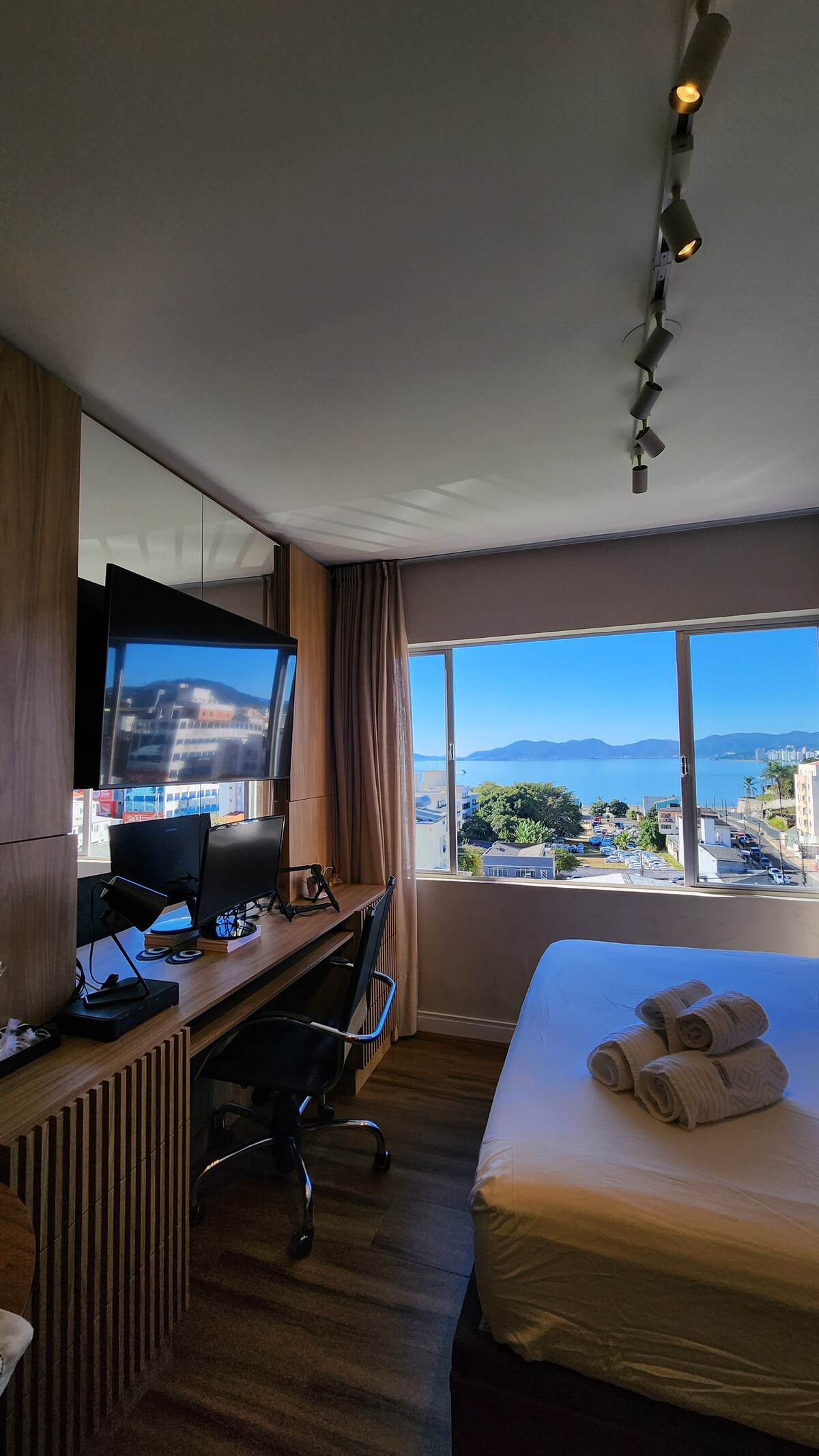 A well-lit bedroom features a spacious bed with neatly arranged towels at the foot. A large window provides a view of the water and city landscape. A desk with two monitors and a chair is situated opposite the bed, enhancing the workspace functionality.