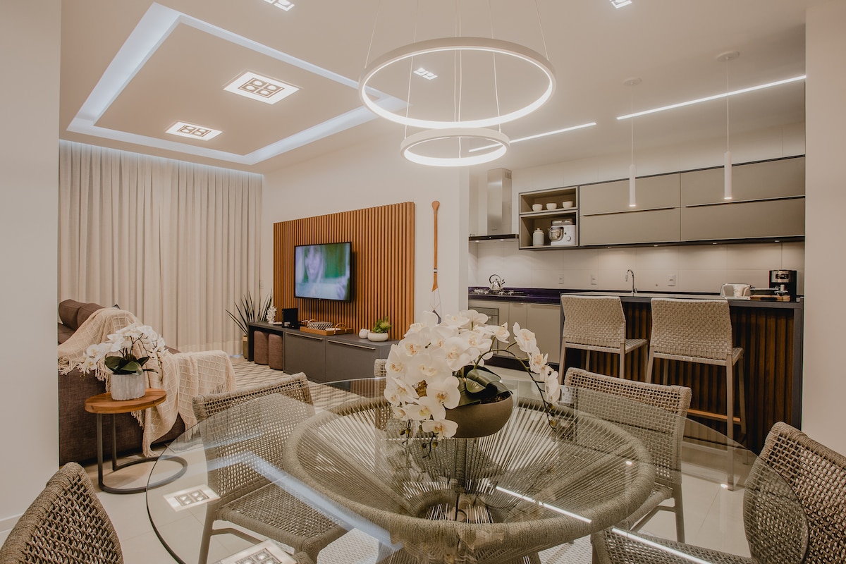 A modern dining area is depicted with a glass table surrounded by woven chairs. A sleek kitchen is visible in the background, showcasing contemporary cabinetry. Soft lighting from a circular fixture enhances the ambiance, while a television is mounted on the wall.