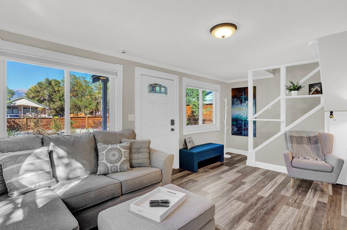 The living area features a spacious gray sectional sofa with decorative pillows, a coffee table, and a comfortable armchair. Large windows allow natural light to fill the space, revealing views of the landscaped yard. A small entryway leads to the staircase and additional seating.