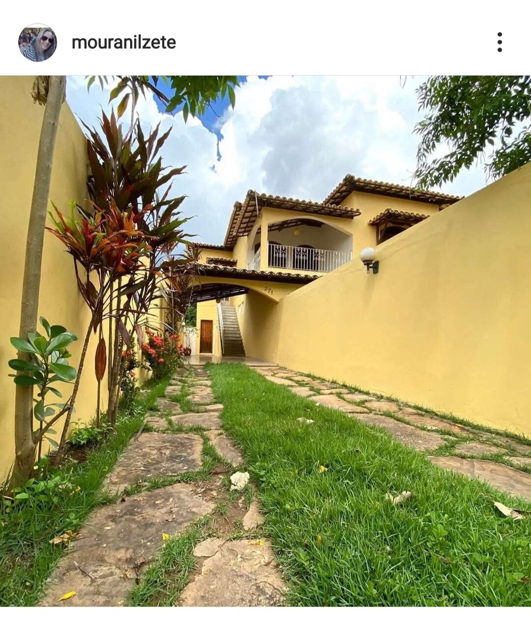 A narrow pathway lined with stone steps leads through a lush green lawn to a two-story yellow house. Vibrant plants are positioned along the sides, while a staircase is visible, guiding the way to the upper level.