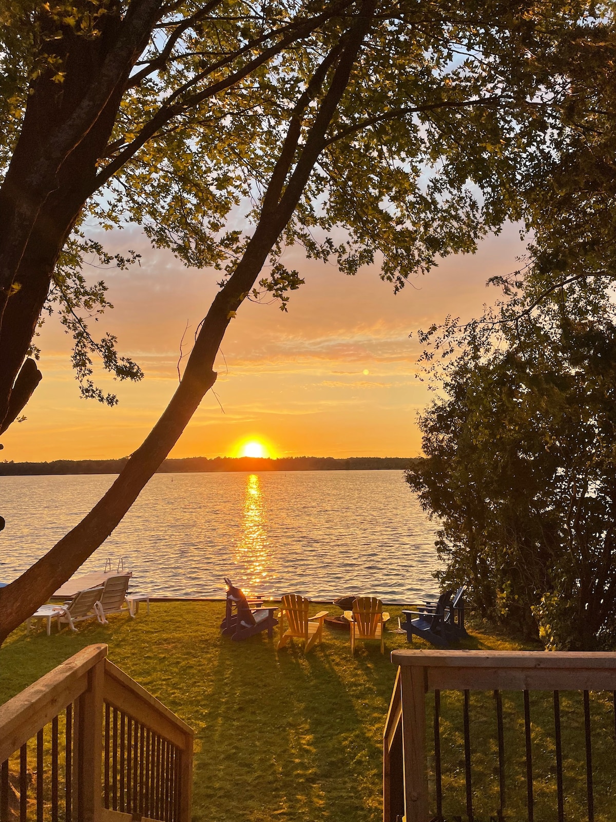 A serene waterfront view captures a sunset over Canal Lake, with golden hues reflecting on the water. Adirondack chairs are placed on the lawn, inviting relaxation, while trees frame the scene, offering a sense of tranquility in the outdoor space.