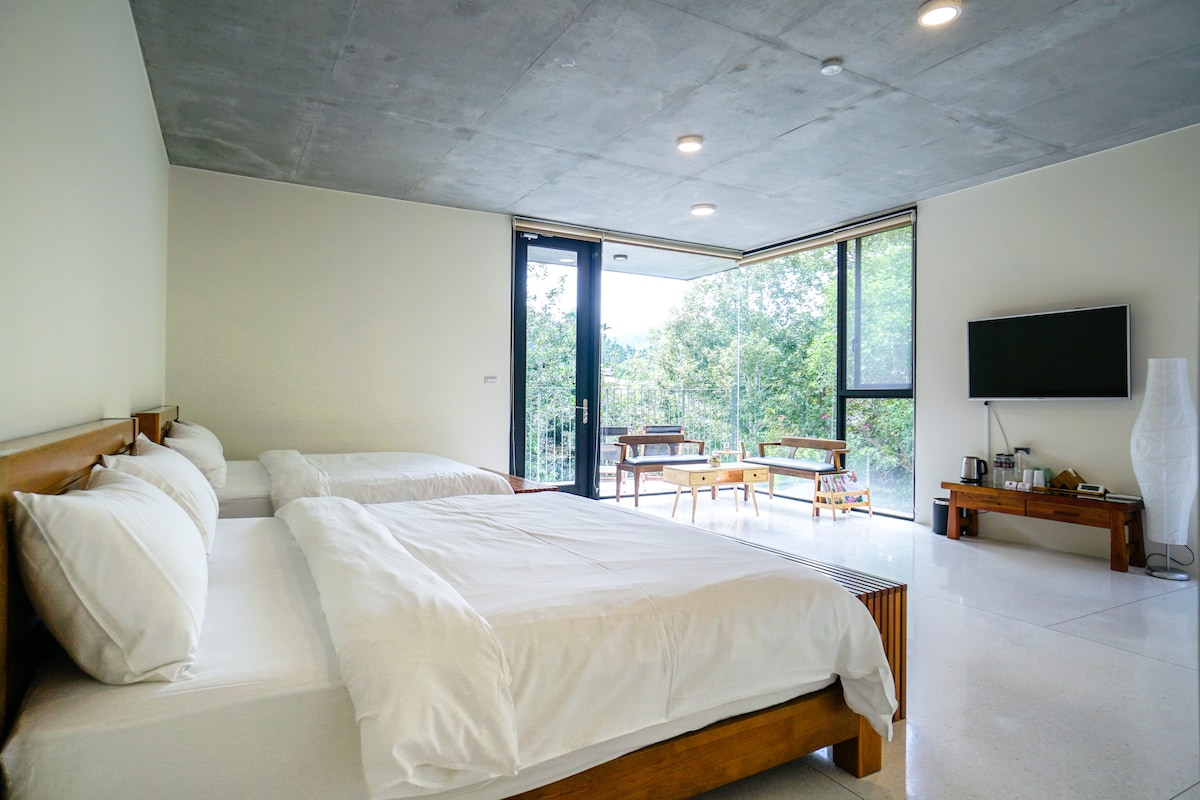A spacious guest room features two large beds with white linens. Large glass doors provide natural light and a view of a green outdoor area. A seating area with wooden furniture is visible, along with a television mounted on the wall.
