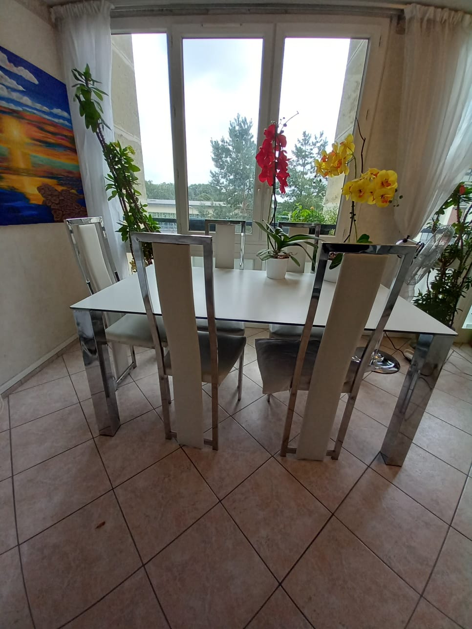 A dining area is seen with a white table surrounded by four modern chairs. Large windows allow natural light to enter, framing views of greenery outside. Indoor plants and colorful flowers add vibrancy to the space, which is punctuated by a wall art piece in bright colors.