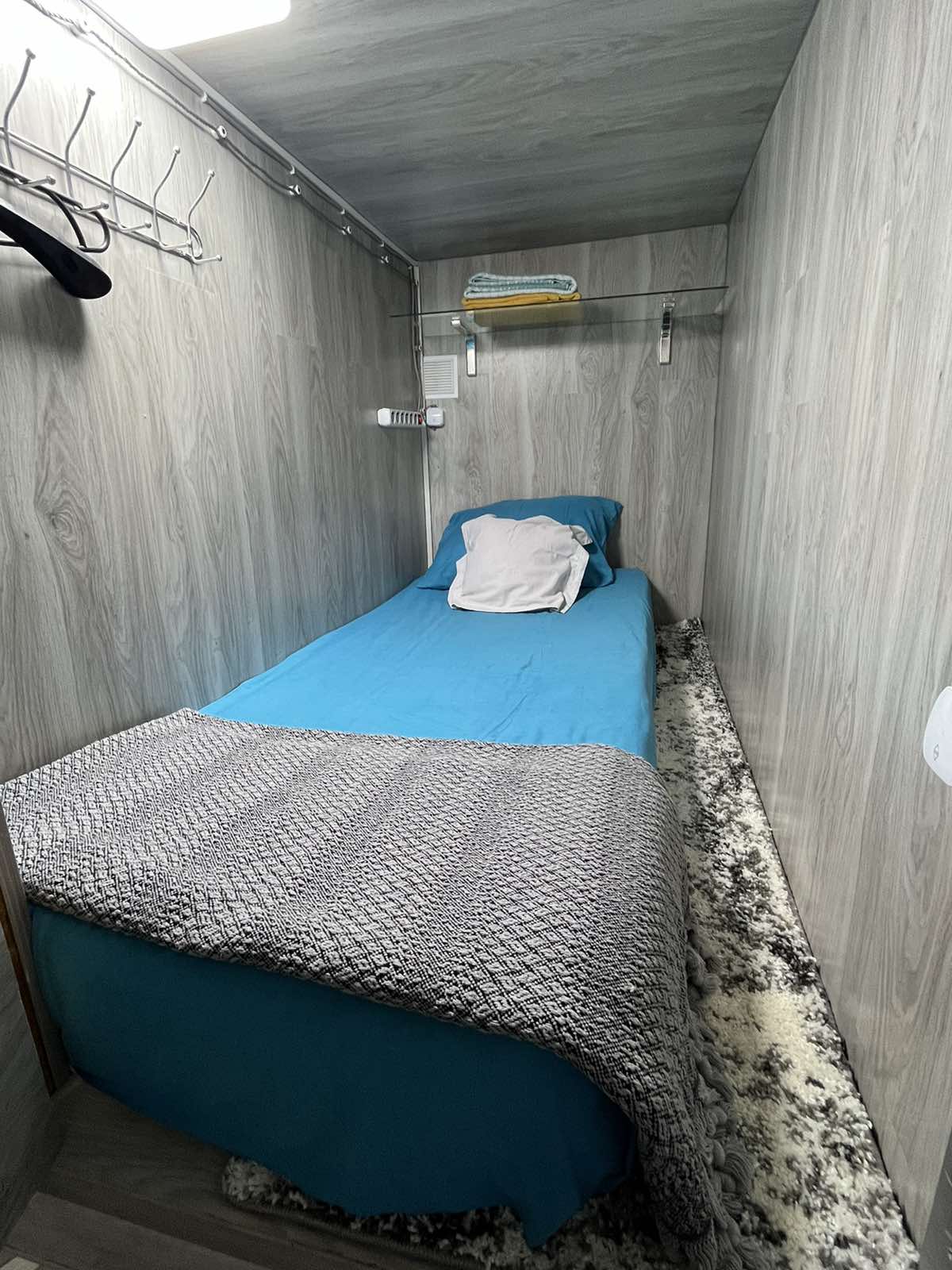 A narrow sleeping area features a single bed dressed in a blue coverlet, complemented by a gray knitted blanket. Light wooden paneling lines the walls, with a small area rug beneath the bed. A shelf above holds towels and a few hangers for clothing.